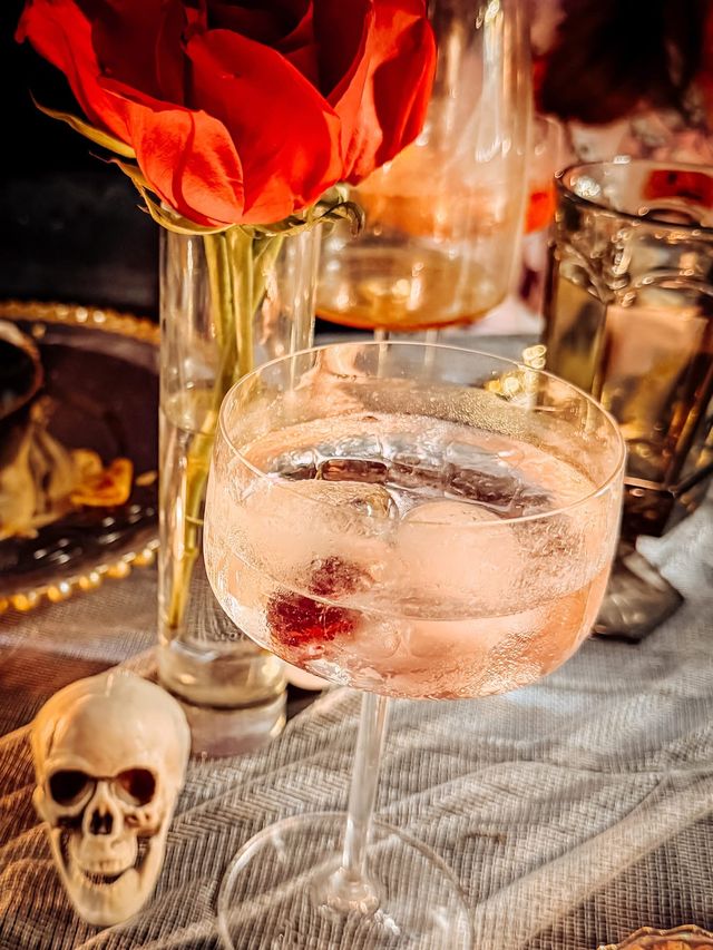A pink cocktail in a coupe glass beside a red rose and a small skull.