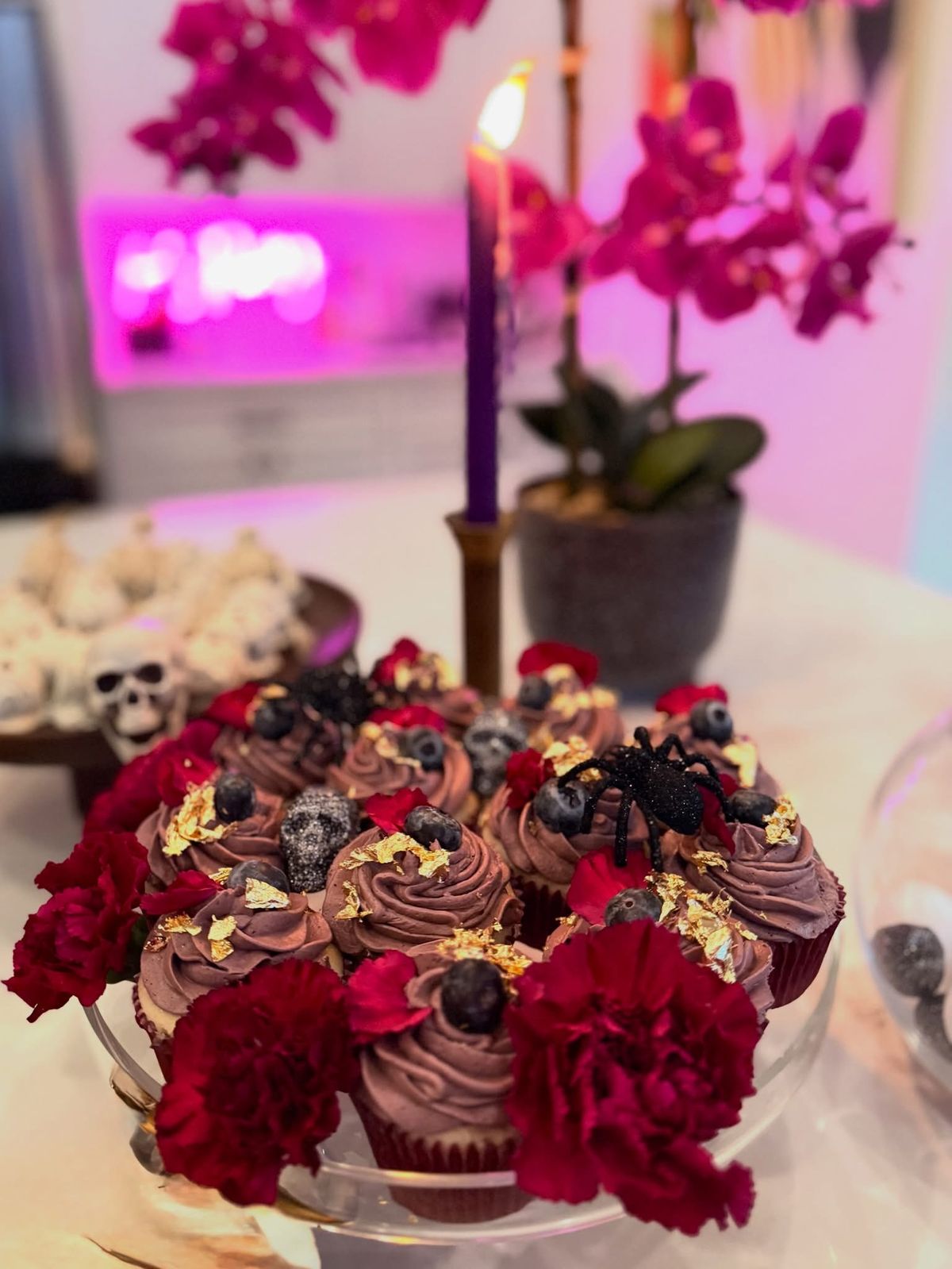 Cupcakes topped with gold leaf and blueberries, ringed with red carnations, with a glitter spider and sugar skulls on top.
