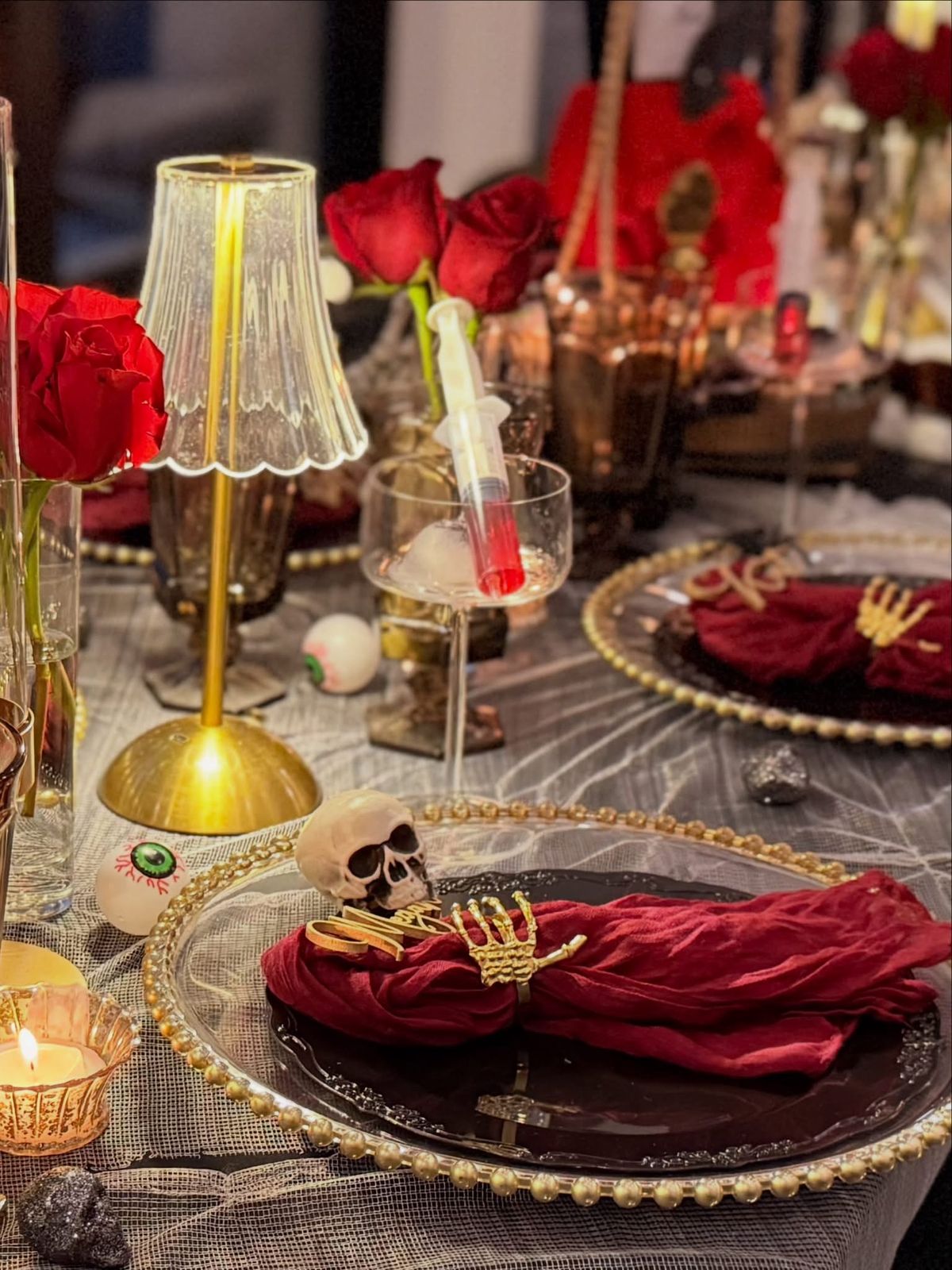 A place setting with a black plate on a gold beaded charger, an oxblood napkin in a gold skeleton-hand ring, a small skull, and a syringe of red liquid resting in a coupe glass.