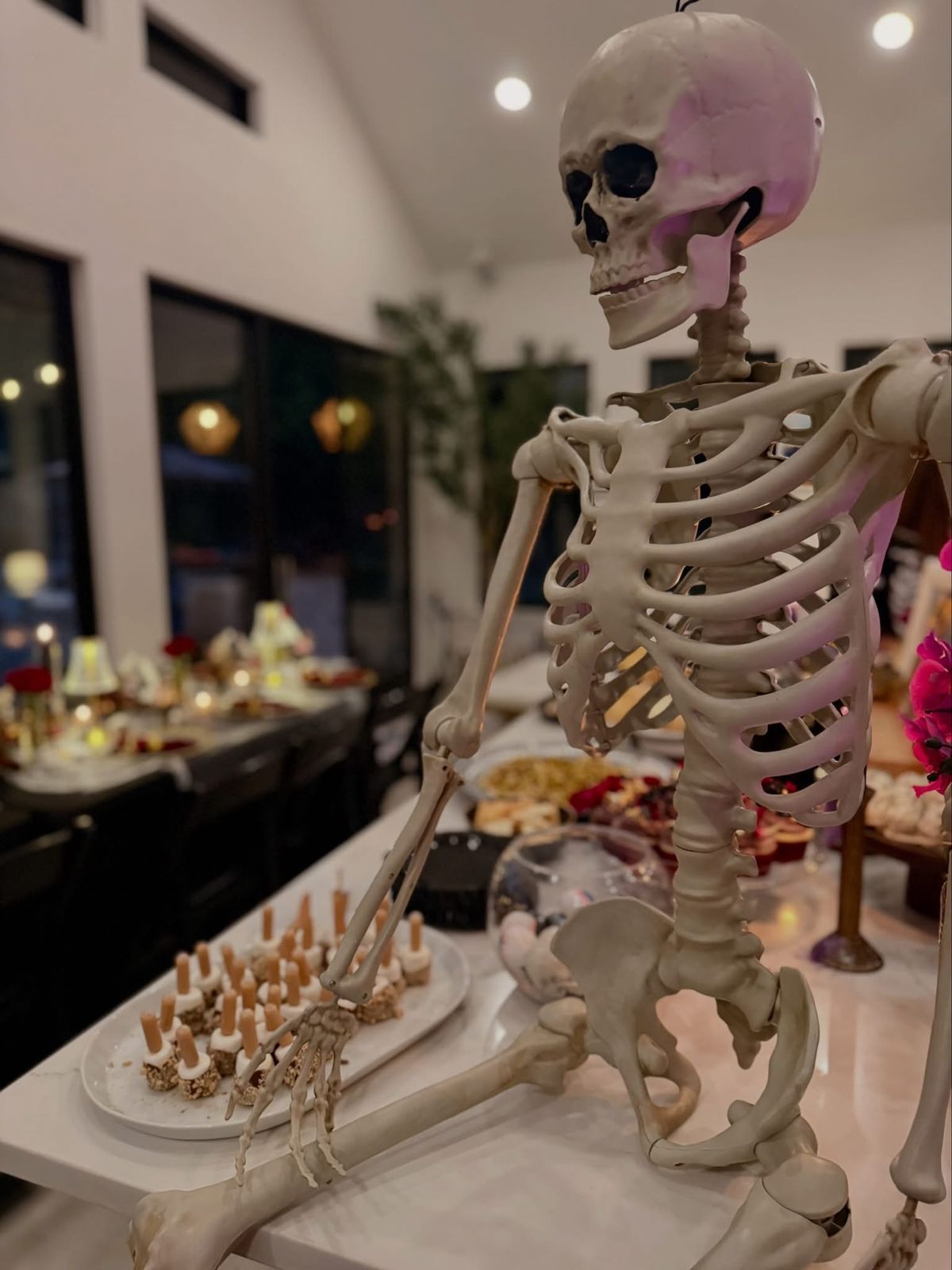 A full-size skeleton seated on the kitchen island next to the food, with the set table glowing in the background.