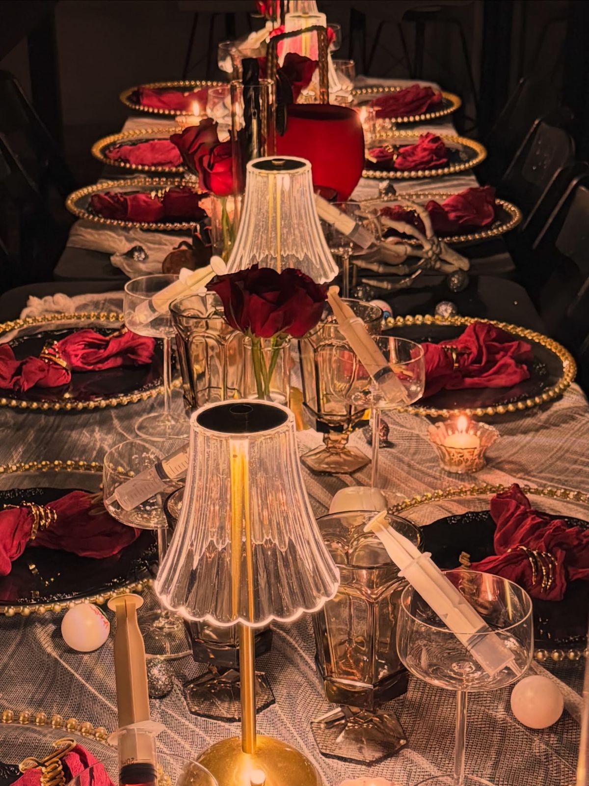 A long candlelit table set for a Halloween dinner: black plates on gold beaded chargers, oxblood napkins, red roses, glass lamps, and coupe glasses down the center.